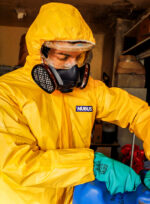 Operario con traje de protección química amarillo Nubus, antiparras de seguridad y guantes verdes de nitrilo DPS, manipulando un bidón azul mientras utiliza la máscara de silicona UCU7502RS con cartuchos de protección instalados.