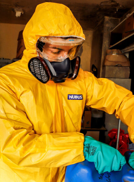 Operario con traje de protección química amarillo Nubus, antiparras de seguridad y guantes verdes de nitrilo DPS, manipulando un bidón azul mientras utiliza la máscara de silicona UCU7502RS con cartuchos de protección instalados.