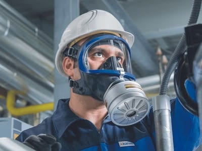 Trabajador en un entorno industrial equipado con casco blanco de seguridad y máscara completa reutilizable Dräger con filtro de rosca, realizando tareas de inspección.